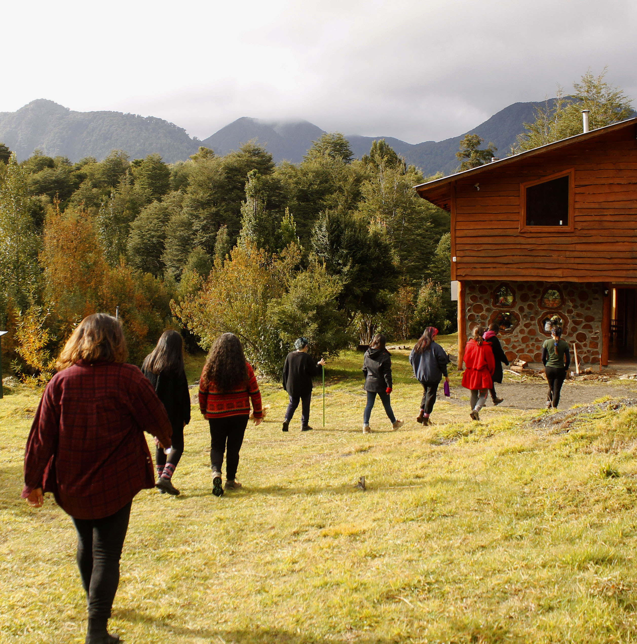 Fotografía en la que en el fondo se observa una casa grande y un gran y frondoso bosque, en el centro se ven nueve mujeres caminando de espalda a distinto paso hacia la casa.