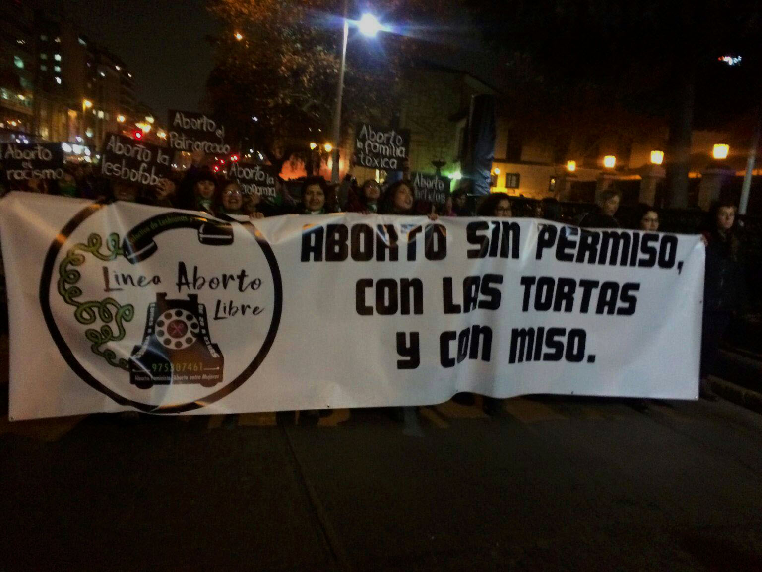 Fotografía de una marcha de noche, se observan muchas mujeres sosteniendo un gran cartel blanco. Al lado derecho del cartel está impreso en grande el logo de la linea aborto libre y a su lado escrito con letras negras y grandes "aborto sin permiso, con las tortas y con miso"